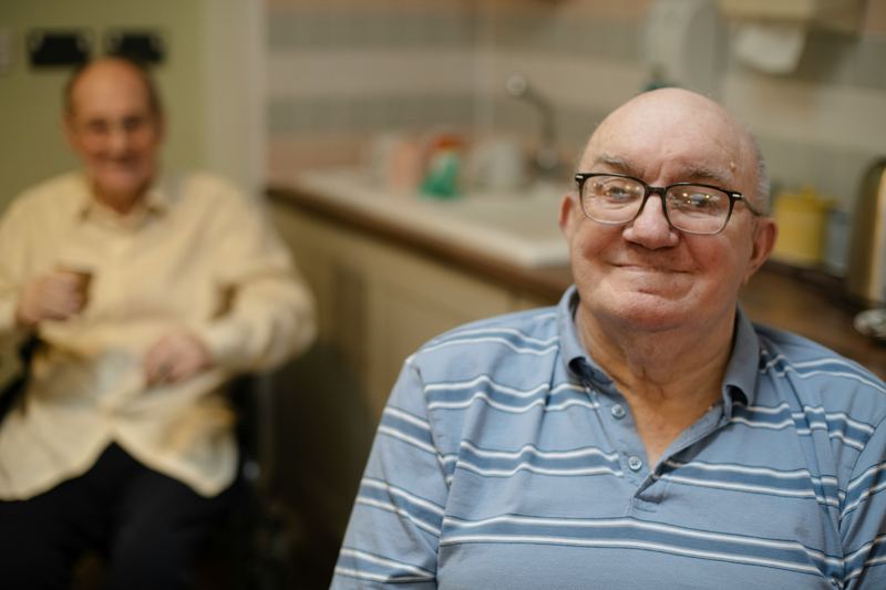 A smiling older man at home, with a friend in the background